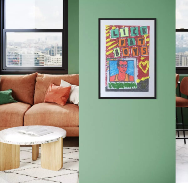 A vibrant outsider art print titled 'LICK FAT BOYS' displayed in a black frame on a sage green wall. The artwork features colorful block letters and a portrait in a pop art style. The print is shown in a modern living room setting with a terracotta velvet sofa, green accent pillows, and a round marble coffee table with wooden legs. The room has large windows with a city view, creating a contemporary urban art collector's space.